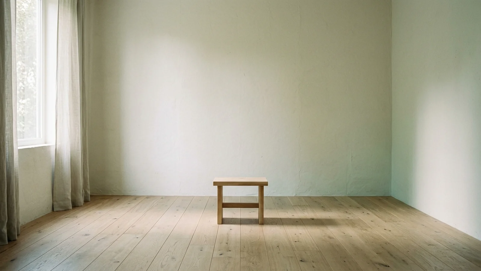 An almost empty room with pale walls, a bare oak floor and one wooden stool in morning light