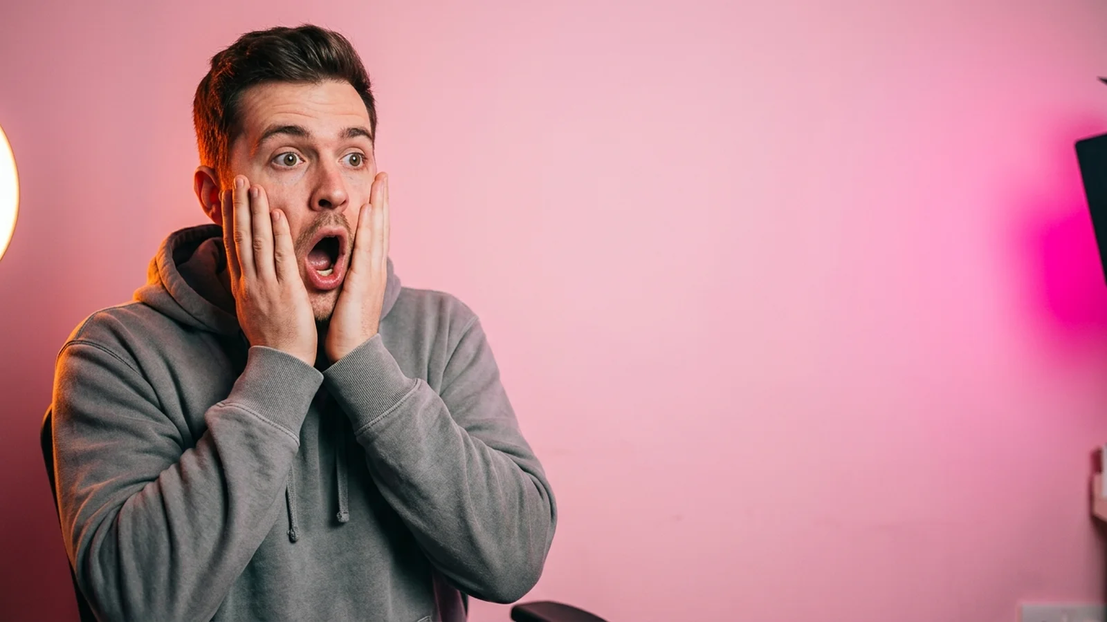 A creator in a grey hoodie with both hands on his cheeks, shocked, in front of a pink wall.